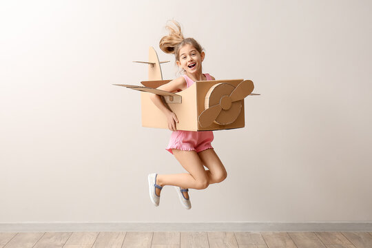 Little girl with cardboard airplane jumping near light wall
