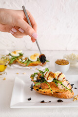 Woman pouring sauce onto tasty bruschetta with eggs and arugula on light background