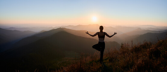 Panoramic view of woman practicing yoga on background of evening mountains. Meditating female is balancing on one leg at sunset. Concept of balance.