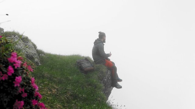 A Closeup Of A Caucasian Male Hiker Sitting At The Edge Of A Cliff, Eating A Bar Of Chocolate With Intense Fog In The Background