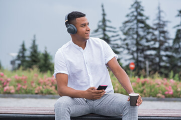 Obraz premium Portrait of a young and happy African American man with headphones. A man sitting on a bench and listening to music, holding a smartphone and a mug of coffee
