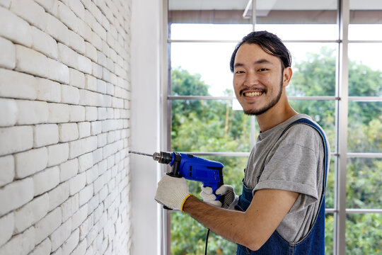 Young Asian Man Using Electric Drill On White Brick Wall In Room, Concept For Home Improvment, DIY Work, Do It Youself