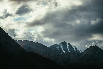 Obraz premium Atmospheric mountain landscape with silhouettes of rocks and snowbound sharp pinnacle under cloudy sky. Great rocks with snow. Beautiful mountain scenery with snow peaked top. Snow-capped pointy peak.