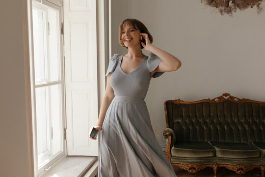 Pretty young lady posing at furnished studio. Cute short-haired brunette wearing maxi dress, smiling and moving with closed eyes near window against vintage sofa