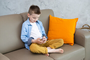 A cute bored boy in a blue shirt sits on the couch at home and uses a mobile phone. The concept of digital addiction in children. Online learning and communication in social networks of children.