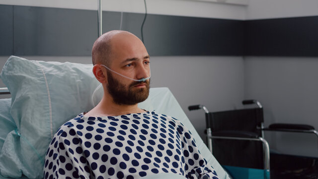 Sad sick man with nasal oxygen tube resting in bad looking into camera during respiratory recovering in hospital ward. Depressed patient waiting for illness breathing treatment