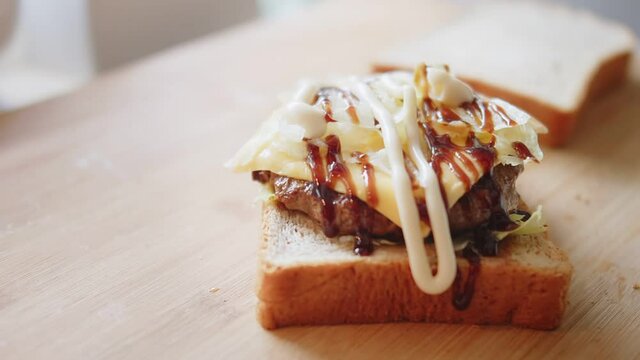 Close up Pouring mayonnaise sauce onto sandwich bread with meat, cheese and vegetables on wooden board. American fast food