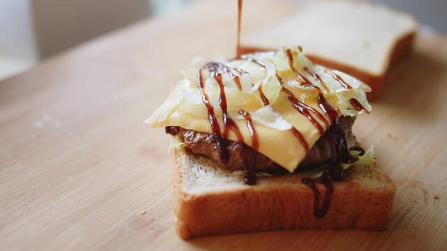 Close up Pouring barbeque ketchup sauce onto sandwich bread with meat, cheese and vegetables on wooden board. American fast food