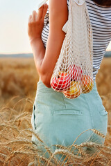 A woman in natural clothes with a string bag made of eco-mesh, fruits, apples is walking along a path in a wheat field.