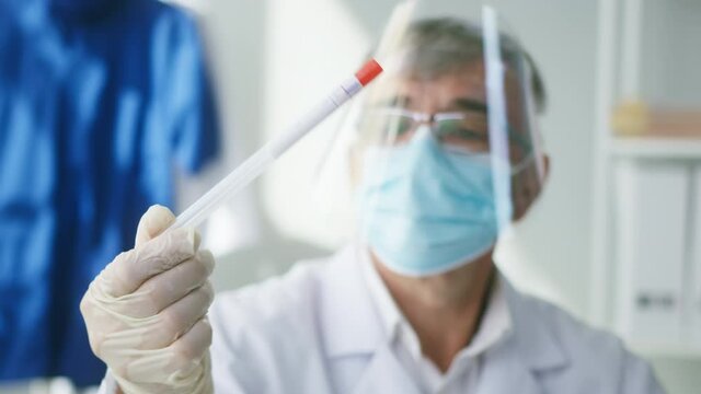 PCR test in laboratory, aged doctor looking at COVID-19 swab collection kit, grey-haired lab worker wearing PPE medical suit, mask, gloves, close-up of test tube for taking OP NP specimen sample