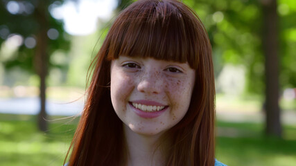 Close up of smiling redhead teen girl looking at camera standing in summer park