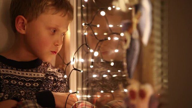 Little boy sitting on windowsill beside bright-glowing Christmas lights waiting for Santa. Holidays, childhood conception.
