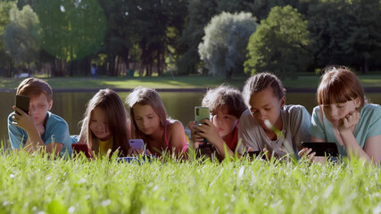 Fototapeta premium Multiethnic teen children using mobile phone lying together on grass in spring park