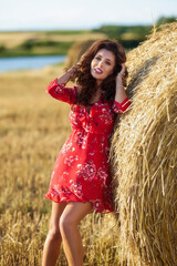 Portrait of beautiful young woman with long hair in floral dress in the summer field near haystacks on sunset.