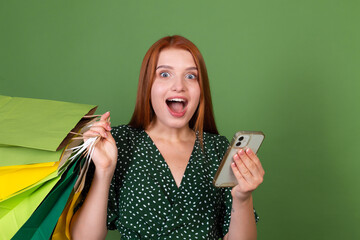 Young red hair woman on green background with  shopping bags and mobile phone shocked amazed