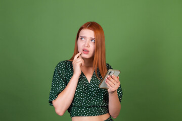 Fototapeta premium Young red hair woman on green background with mobile phone confused thoughtful look aside