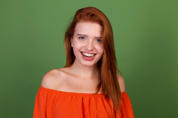 Young red hair woman in casual orange blouse on green background look to camera excited smile laugh