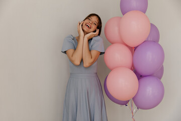 Lifestyle portrait of young lady near pastel pink and violet balloons. Adorable dark-haired girl with narrowed eyes, blue dress, touching face and smiling against light background