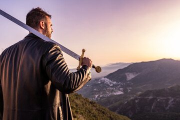 man dressed in a leather trench coat with a large sword resting on his shoulder at sunset with mountains and a village in the background