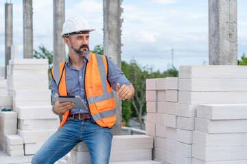 Foreman wearing an engineer helmet holding a tablet inspects construction work at a construction site 