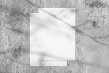 Two empty white rectangle poster or card mockups lying on top of each other with soft tree leaves and branches shadows on neutral light grey concrete wall background. Flat lay, top view
