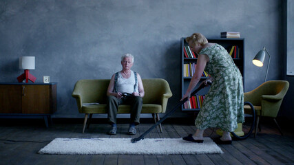 Elderly woman vacuuming the floor with husband sitting on couch