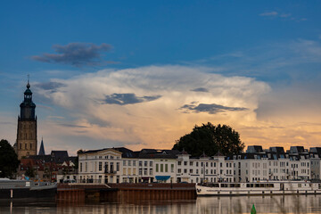 Towering cumulonimbus rain cloud above cityscape of Zutphen in The Netherlands at sunset with orange colors contrasted against a blue sky. Dutch climate and weather condition landscape.