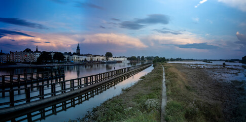 Panorama showing floodplains and cityscape of Zutphen in The Netherlands with large cumulonimbus rain clouds above in colorful sunset tones. Dutch climate weather condition landscape.