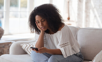 Thoughtful young mixed raced woman using smartphone enjoying chatting with boyfriend on phone sitting on sofa in boho interior. The concept of a long-distance online romantic relationship and dating.