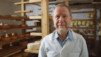 Adult bearded blonde cow farmer smiling looking at camera with cheese rounds on background. Eco...