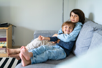 Two brothers are sitting on a sofa at home. The elder boy hugs the younger. Siblings good relations. © Anna