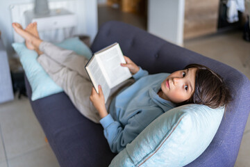 Young boy wearing light blue sweatshirt, lying on  the sofa, with open book in front of him, looking at the camera.