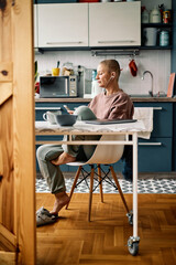 An attractive caucasian senior woman is sitting on a chair in her kitchen in the morning and listening to music on the phone of having a video call with loved ones.