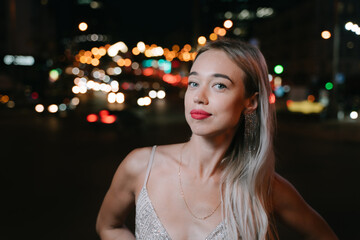 Big city night life. Beautiful young blonde woman with red lips and blue eyes looking at camera. fancy golden cocktail Glided dress and long earrings. Summer party mood. Bokeh lights background. 