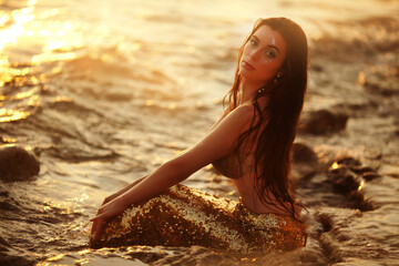 Beautiful fashionable mermaid with red hair holding shell sitting on rock near sea