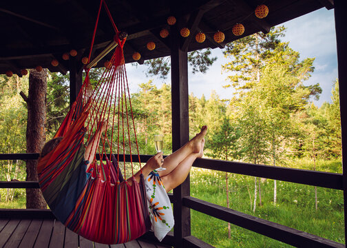 Young woman enjoy alone time in forest resort in Nordic pine tree forest in summer, relax sitting in swing hanging textile chair on balcony, drinking mulled wine.