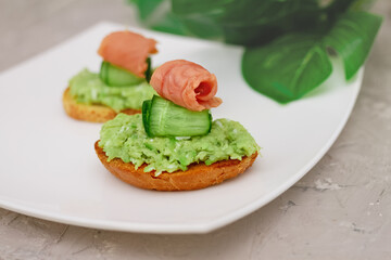 sandwich with guacamole, cucumber and red fish on white plate on concrete background with monstera leaves