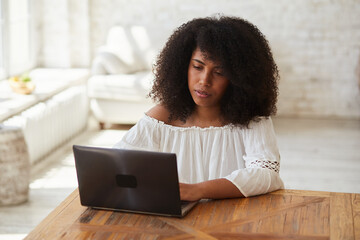 Thoughtful black female blogger using laptop for writing posts in social network. Professional busy mixed race freelancer sitting on coach and search for inspiration and solving problems.