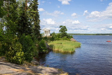 Fototapeta na wymiar VYBORG, Leningrad REGION, RUSSIA-JULY, 19, 2021: view of the white temple of Neptune and the green foliage of trees on a summer cloudy day in the Monrepos Park and the beautiful shore and blue water