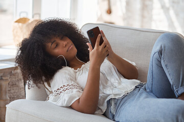 Happy african woman in headphones looking at smartphone, enjoying video meeting with friends and colleagues, online virtual conversation. Female black student sitting on sofa and making video call. - Powered by Adobe