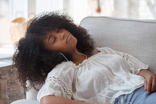 A relaxed young black smiling woman wearing headphones, resting on a comfortable couch, listening music tracks, audiobooks or podcasts and enjoying weekend stress-free leisure time indoors.