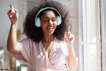 Happy hilarious black multiracial lady wearing headphones near the window listening to modern hip hop singing song with eyes closed resting in comfortable apartment at home