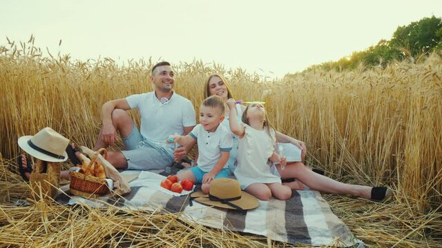 Picnic with soap bubbles in wheat field at harvest season. Little children blowing bubbles and parents watching them. Happy family spending time together. Concept of fun