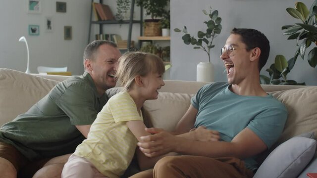 Two men spending time with their little daughter having fun in living room tickling each other, slow motion shot