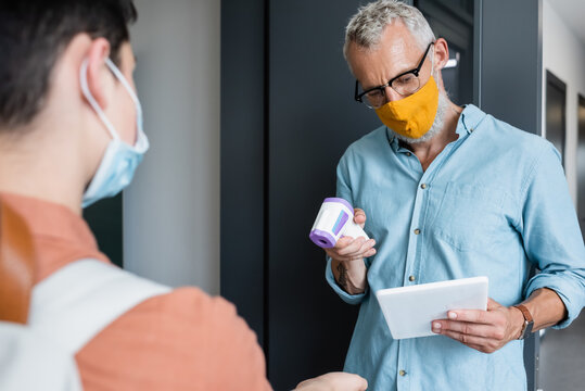 mature teacher in orange medical mask holding pyrometer while looking at digital tablet near blurred boy