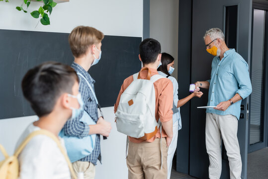 mature teacher in medical mask checking temperature of teenage pupils with pyrometer