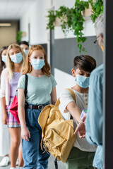 blurred teacher checking temperature of multiethnic pupils in medical masks with pyrometer