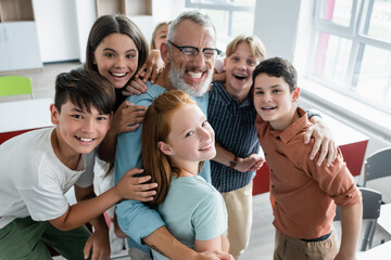 joyful multiethnic classmates looking at camera while hugging happy teacher laughing with closed...