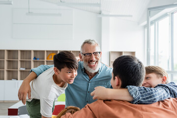 Fototapeta premium cheerful multiethnic boys embracing laughing middle aged teacher in classroom