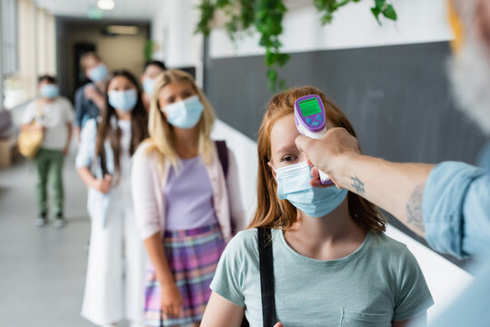 selective focus of pyrometer in hand of teacher checking temperature of pupils standing in line in medical masks